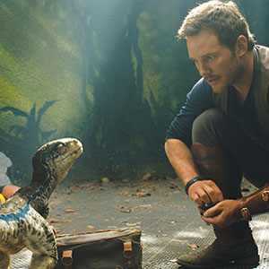A man crouches down and looks attentively at a small, blue-and-white dinosaur in a jungle-like setting, with a closed brown bag on the ground between them. Sunlight filters through the trees in the background.