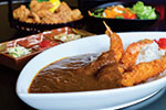 A white oval plate of Japanese curry with rice and breaded fried shrimp. In the background, there is a plate of fried food and a bowl of salad with colorful vegetables.