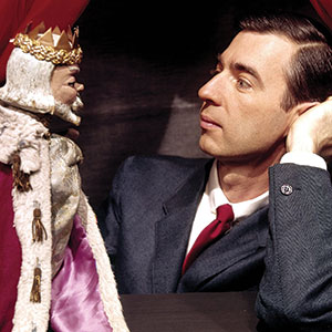 A man in a suit and red tie smiles gently at a puppet wearing a crown and royal robe. The man is resting his head on his hand, and the puppet faces him under a red curtain.