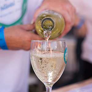 A close-up of a person pouring white wine from a bottle into a wine glass. The wine is bubbly, and the person is wearing a blue wristband and a white shirt. The background is blurred.