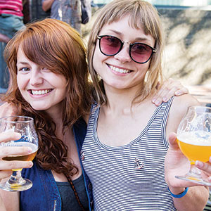 Two smiling women holding glasses of beer pose together outdoors. One has long red hair and the other wears sunglasses and a striped tank top. They appear happy and are enjoying a sunny day.