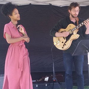 A woman in a pink dress sings into a microphone while a man beside her plays an electric guitar on stage, both performing under a canopy with a black backdrop.