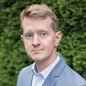 A man with short light brown hair, wearing a light blue blazer and a white collared shirt, stands outdoors in front of a background of green foliage, looking directly at the camera with a neutral expression.