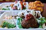 Close-up of takeout containers filled with Mediterranean food, including falafel topped with sauce, tabbouleh salad, waffle fries, and other assorted sides, served on a bed of greens.