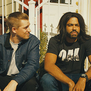 Two men sit on outdoor steps in front of a white metal gate. The man on the left has short, light brown hair and wears a blue jacket, while the man on the right has long dark hair and wears a black T-shirt.