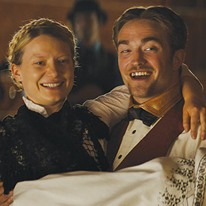 Two people in vintage clothing smile and dance closely together. The woman wears a black lace dress, and the man wears a bow tie and vest, holding her with one arm. A blurred figure is visible in the background.