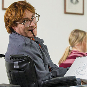 A man with red hair and glasses sits in a wheelchair, holding a pen in his mouth and drawing in a sketchbook. He looks back and smiles. A girl with blonde hair and a striped shirt is sitting in the background.