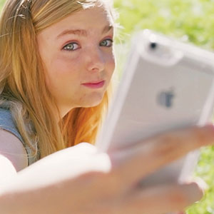 A young girl with long blonde hair holds a smartphone in front of her, taking a selfie outdoors with green grass in the background.