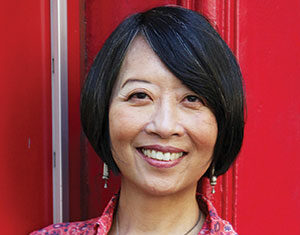 A woman with short black hair and a floral patterned red shirt smiles while standing in front of a bright red background. She is wearing dangling earrings and a silver necklace.