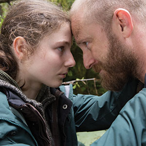 A young woman and a bearded man touch foreheads closely, looking into each others eyes with serious expressions. They are outdoors, wearing jackets, surrounded by greenery.
