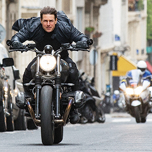 A man in a black jacket rides a motorcycle at high speed down a city street, leaning forward intensely, with a police officer on a motorcycle following in the background.