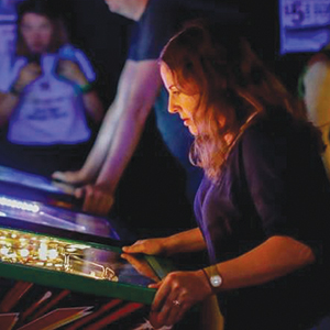 A woman intently plays a pinball machine in a dimly lit arcade, with colorful lights illuminating her face while others watch in the background.