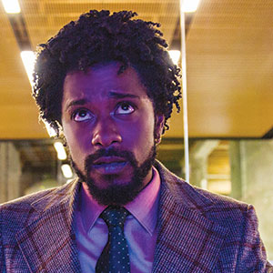 A man with curly hair and a beard, wearing a checkered suit jacket, shirt, and tie, looks upward with a serious expression. The lighting is purple, and the background features a modern interior with bright ceiling lights.