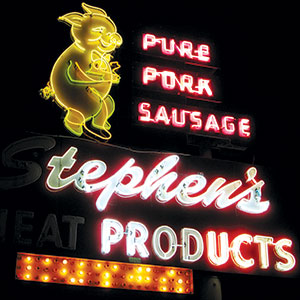 A neon sign at night featuring a smiling pig holding sausages, with glowing red and white text that reads PURE PORK SAUSAGE and Stephens PRODUCTS.