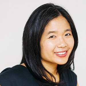 A woman with straight, shoulder-length black hair and a black top smiles at the camera against a plain light background.
