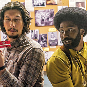 Two men sit side by side in front of a bulletin board with photos. One, with long hair and a plaid shirt, examines a red card, while the other, in a yellow shirt, looks at him with curiosity.