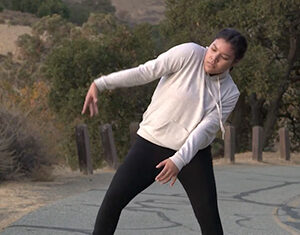 A person wearing a light hoodie and black pants stands barefoot on a paved road outdoors, bending their knees and leaning sideways with one arm extended and the other bent, as if dancing or posing. Trees and hills are in the background.