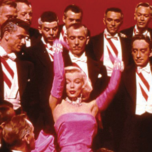 A blonde woman in a pink dress and long pink gloves raises her arms while surrounded by men in tuxedos with red and white sashes, against a red background.