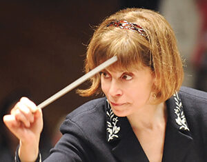 A woman with short brown hair and a dark jacket decorated with white embroidery conducts an orchestra, holding a baton in her raised right hand and looking intently forward.