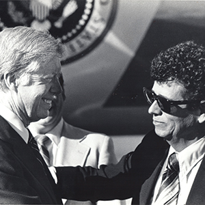 Two men in suits greet each other warmly outdoors. One, smiling, has light hair; the other wears sunglasses and has curly dark hair. A partial U.S. presidential seal is visible in the background.