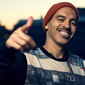 A man wearing a red beanie and a patterned sweater smiles warmly and points toward the camera, with an urban background blurred behind him at sunset.