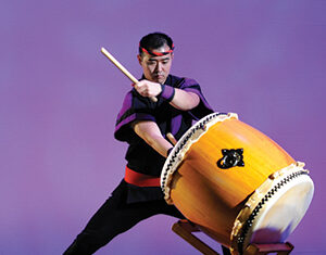 A man wearing traditional attire energetically plays a large taiko drum with wooden drumsticks on a stand, against a purple background.