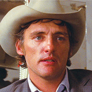 A man wearing a light-colored cowboy hat and a gray jacket looks ahead with a serious expression. His hair is slightly curly, and the background is blurred with some lights visible.