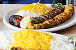 A plate of Persian kebab with saffron rice, grilled tomato, a wedge of lemon, and herbs. Another plate of rice with saffron is visible in the foreground.