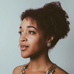 A young woman with natural curly hair styled up, wearing a decorative earring and a chunky beaded necklace, looks thoughtfully to the side against a light gray background.