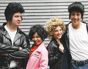 Four people dressed in Grease-inspired costumes, with two men in black leather jackets and wigs, and two women—one in a pink jacket with Rizzo on it and curly hair, the other in a black jacket and blonde curly wig—smiling together.