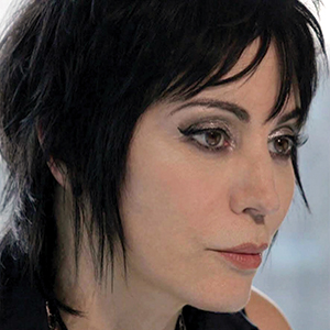 A person with short, layered black hair and dramatic eye makeup looks thoughtfully to the side. The background is softly blurred, keeping the focus on their face and expression.