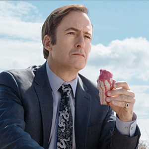 A man in a suit sits outdoors under a blue sky, holding a cup of ice cream with red syrup. He looks into the distance with a serious expression.