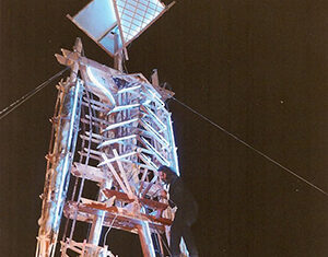 A person climbs a large wooden sculpture shaped like a human figure, illuminated by lights at night, with wires anchoring the structure for support.