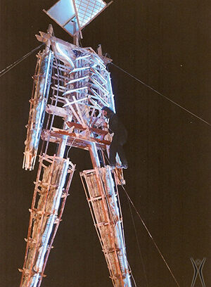 A person climbs a large wooden sculpture shaped like a human figure, illuminated by lights at night, with wires anchoring the structure for support.
