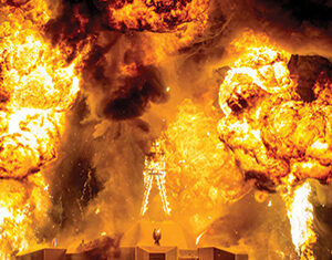 Silhouetted people stand in front of a large wooden structure engulfed in flames, with massive fireballs and thick smoke erupting into the night sky.