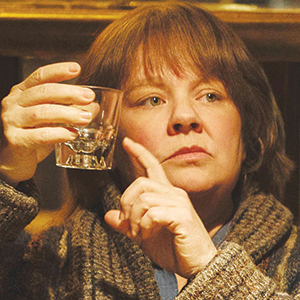 A woman with brown hair and a serious expression holds up an empty glass, inspecting it closely. She is wearing a knit sweater and sitting indoors with a blurred background.