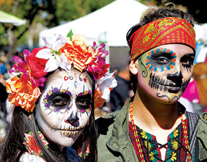 Two people with colorful Day of the Dead face paint and festive attire; one wears a floral headpiece and white dress, the other a red headband and green jacket with beaded patterns, both posing outdoors at a celebration.