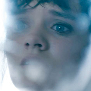 Close-up of a persons face with blue eyes, looking worried or scared, viewed through a frosted or foggy surface that blurs parts of their features.