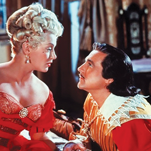 A woman in an elaborate red dress with blonde curled hair looks intently at a man in a period costume with dark hair, who is kneeling and looking up at her. The scene appears dramatic and set in a historical era.