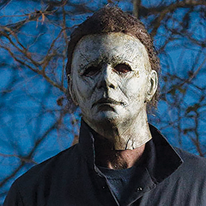 A person wearing a pale, expressionless mask with dark eye holes and messy hair, dressed in a dark outfit, stands outdoors with bare tree branches in the background.