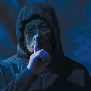 A person wearing a dark hooded sweatshirt and a rough, textured mask holds a finger to their lips, signaling for silence, with a dark, blue-lit background.