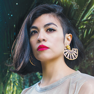 A person with straight, dark shoulder-length hair and bold pink lipstick poses outdoors. They are wearing large, geometric gold earrings and a sheer, light-colored top. Green foliage is visible in the blurred background.