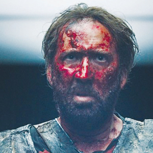 A man with a bloodied face and messy hair stares ahead with a serious expression, wearing rugged clothing, illuminated by dim lighting from above.