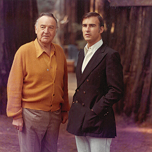 Two men stand outdoors in front of tall trees. The older man on the left wears a mustard cardigan and striped pants; the younger man on the right wears a dark blazer and light pants. Both look toward the camera.
