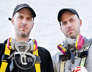 Two men wearing baseball caps, safety harnesses, and respirator masks stand side by side in front of a white wall, looking at the camera. One man holds a paint-splattered glove and a spray can.
