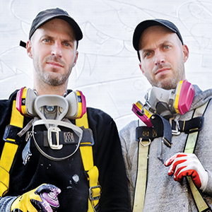 Two men wearing baseball caps, safety harnesses, and respirator masks stand side by side in front of a white wall, looking at the camera. One man holds a paint-splattered glove and a spray can.
