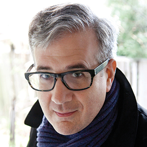 A man with short gray hair and thick black glasses looks at the camera. He is wearing a dark coat and a navy blue scarf. The background is softly blurred with hints of greenery and a fence.