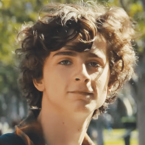 A young person with curly brown hair and light skin is outdoors, standing in front of blurred green trees on a sunny day.