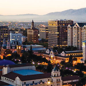 A cityscape at dusk featuring tall buildings, historic architecture, and glowing lights, set against distant mountains under a fading sky.