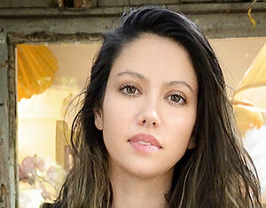 A woman with long, wavy brown hair and light skin looks directly at the camera with a neutral expression. She is standing in front of a window with soft, warm lighting and blurry objects in the background.